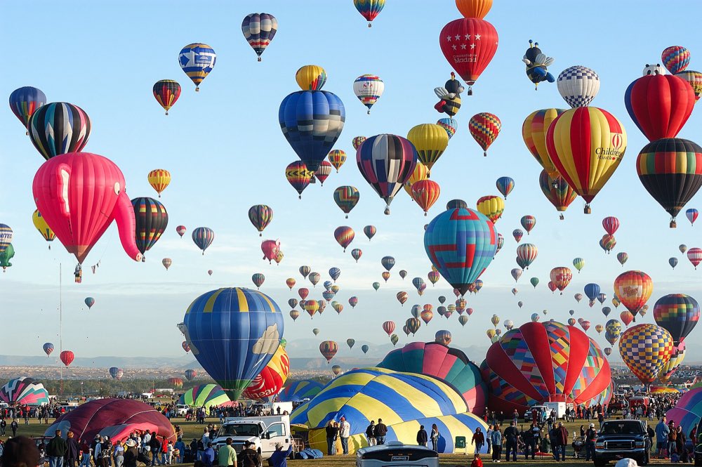 Dozens of brightly striped hot air balloons rising into a clear blue morning sky over the Balloon Fiesta launch field, with more balloons still inflating on the grass