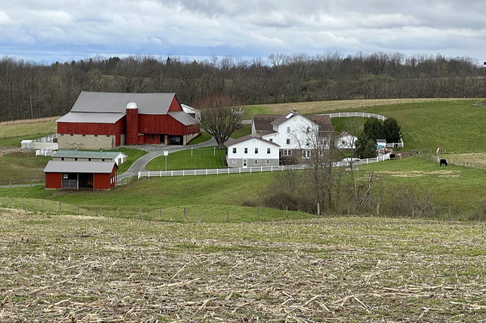 A red barn with a silo, white farmhouses and white board fences on green rolling hills under a grey sky, with a stubble field in the foreground