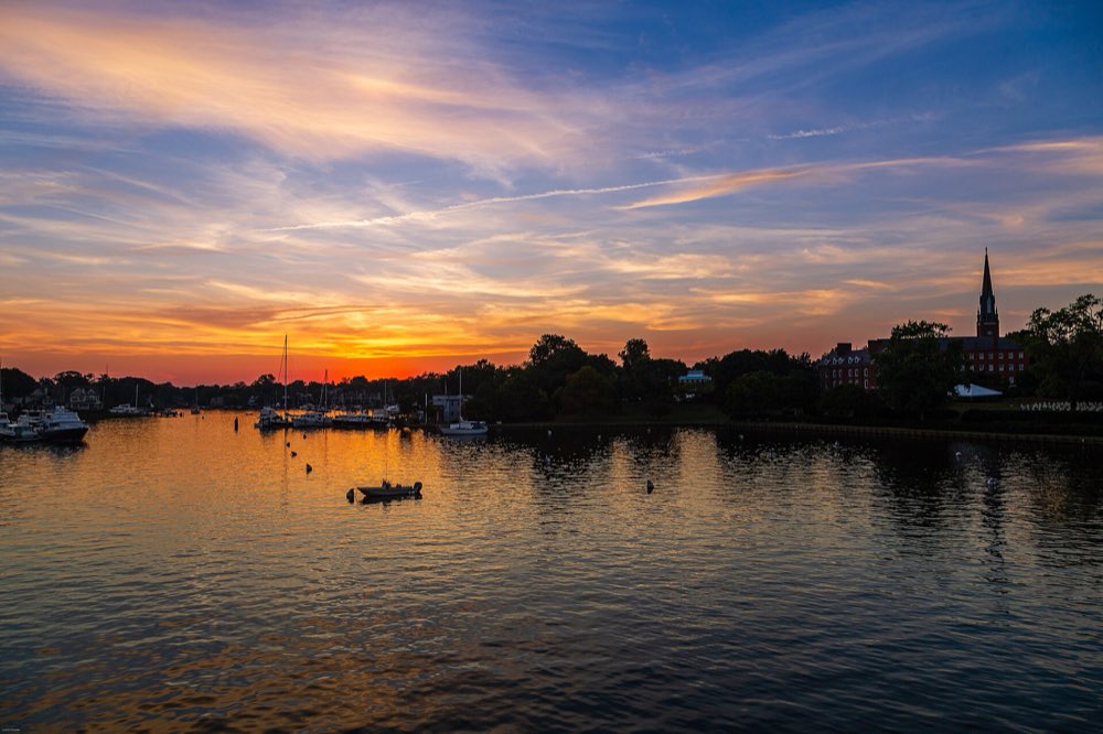 Sunset over a calm Annapolis harbor, with moored boats and sailboat masts along the shore, a church spire against an orange and blue sky, and a small boat in the foreground water