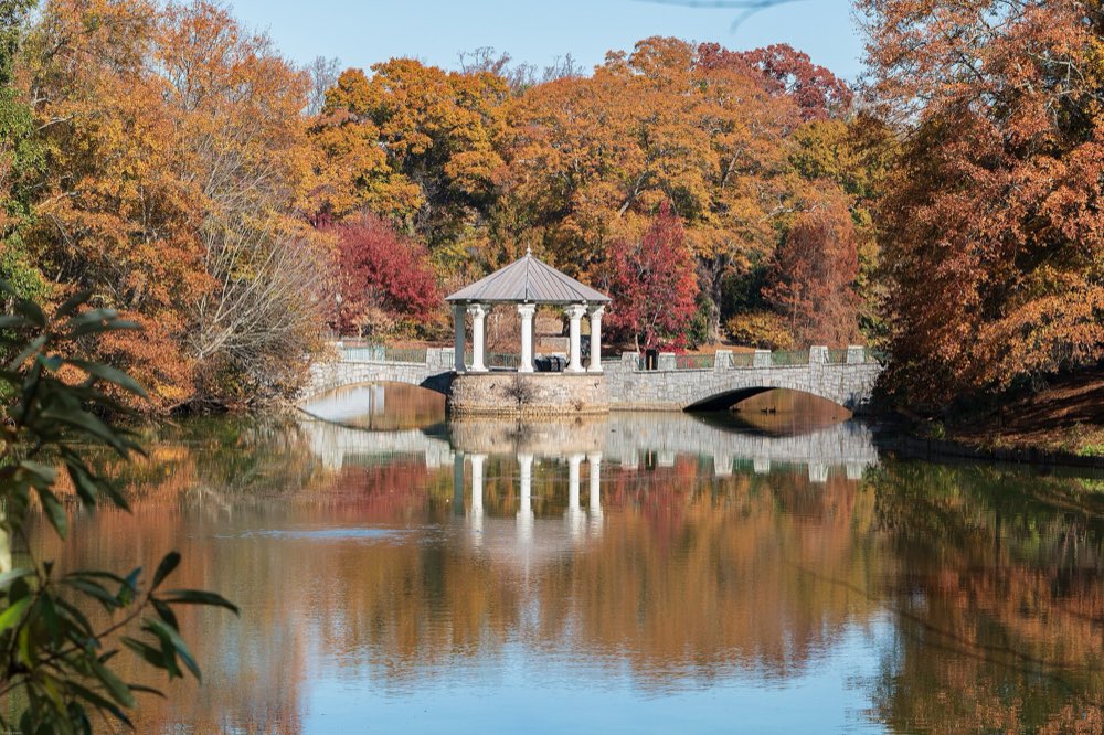 A white gazebo and stone bridge on the edge of Lake Clara Meer in Piedmont Park, framed by orange and red autumn trees and reflected in the still water