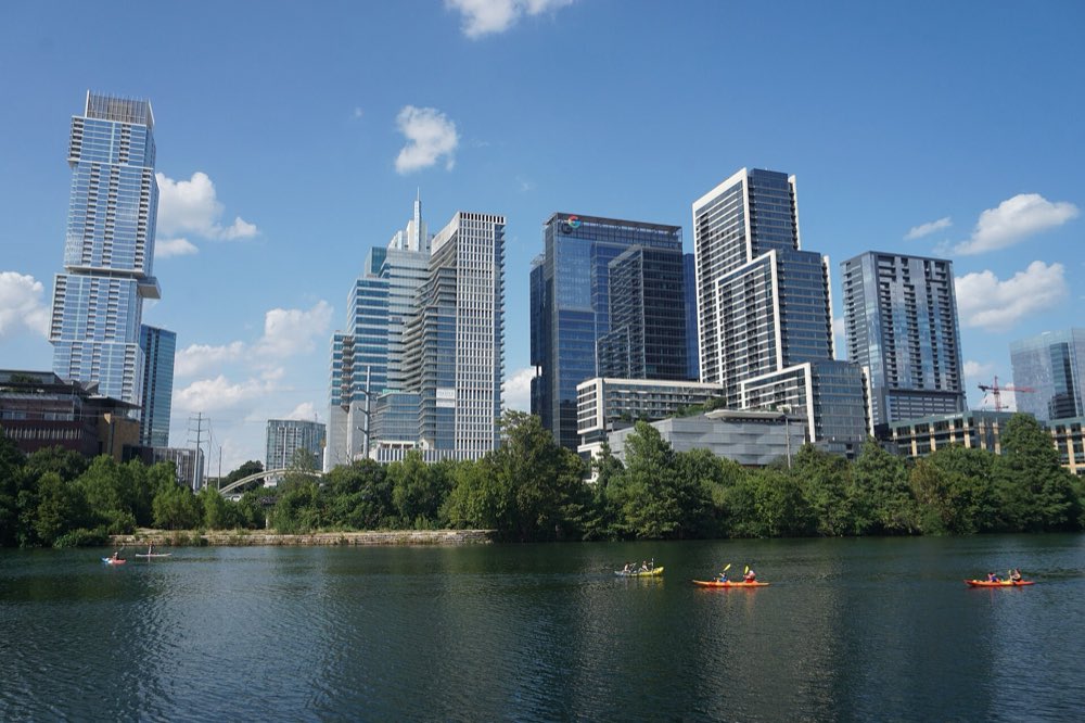 Kayakers paddling across Lady Bird Lake below a row of downtown Austin towers and a tree-lined shore under a blue sky