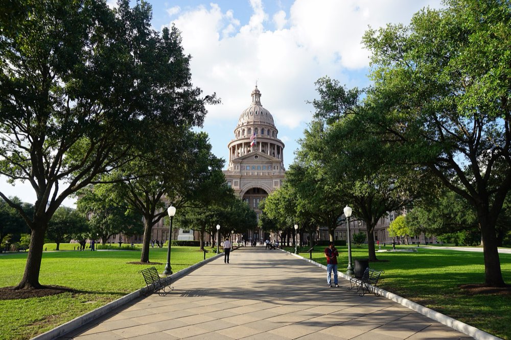 A wide paved walk lined with live oaks and lawns leading to the domed Texas State Capitol under a blue sky with white clouds