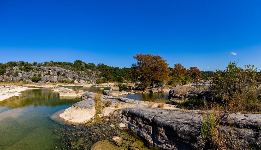 The Pedernales River spreading in clear pools between pale limestone ledges, with bald cypress trees turning rust-orange under a deep blue sky