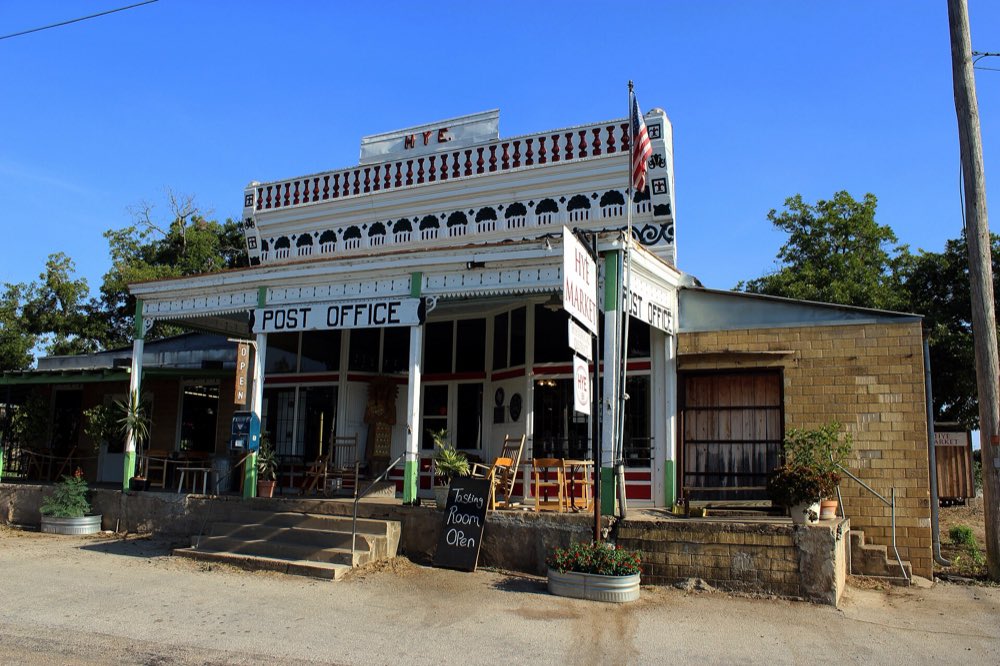 The old Hye General Store and Post Office, a white wooden storefront with an ornate railing, a flagpole and potted plants under a clear blue sky