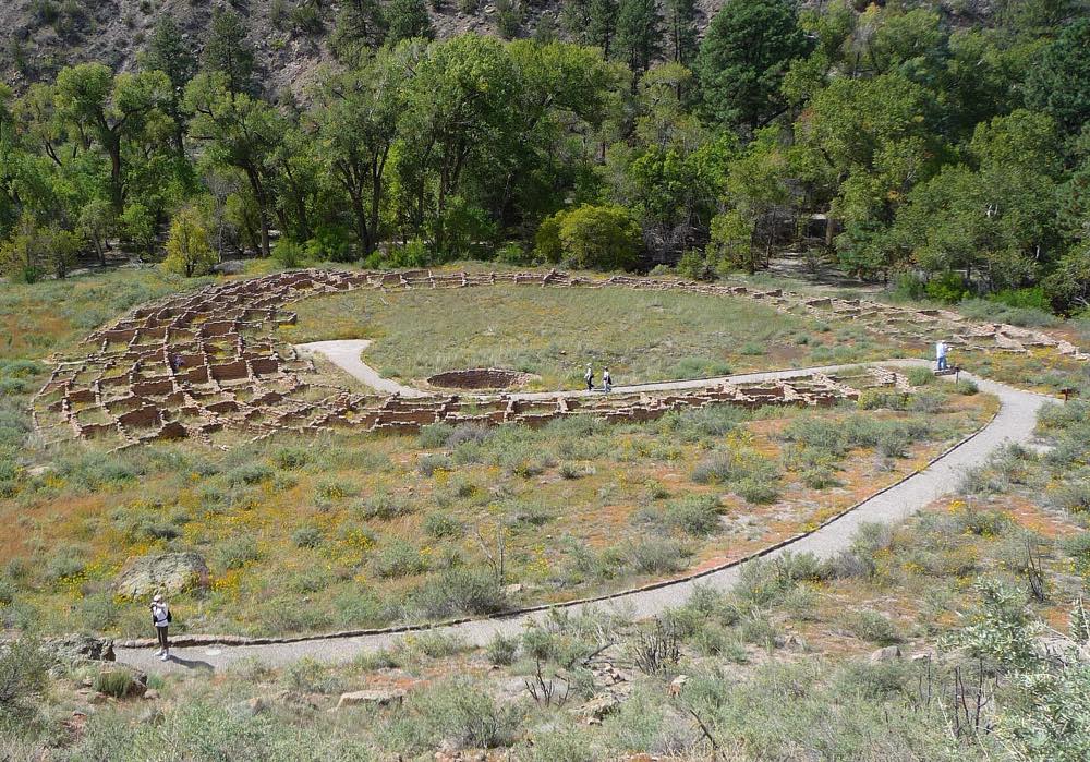 The round stone foundations of Tyuonyi pueblo on the floor of Frijoles Canyon, seen from the cliff trail above, with a paved path curving past and green cottonwoods behind