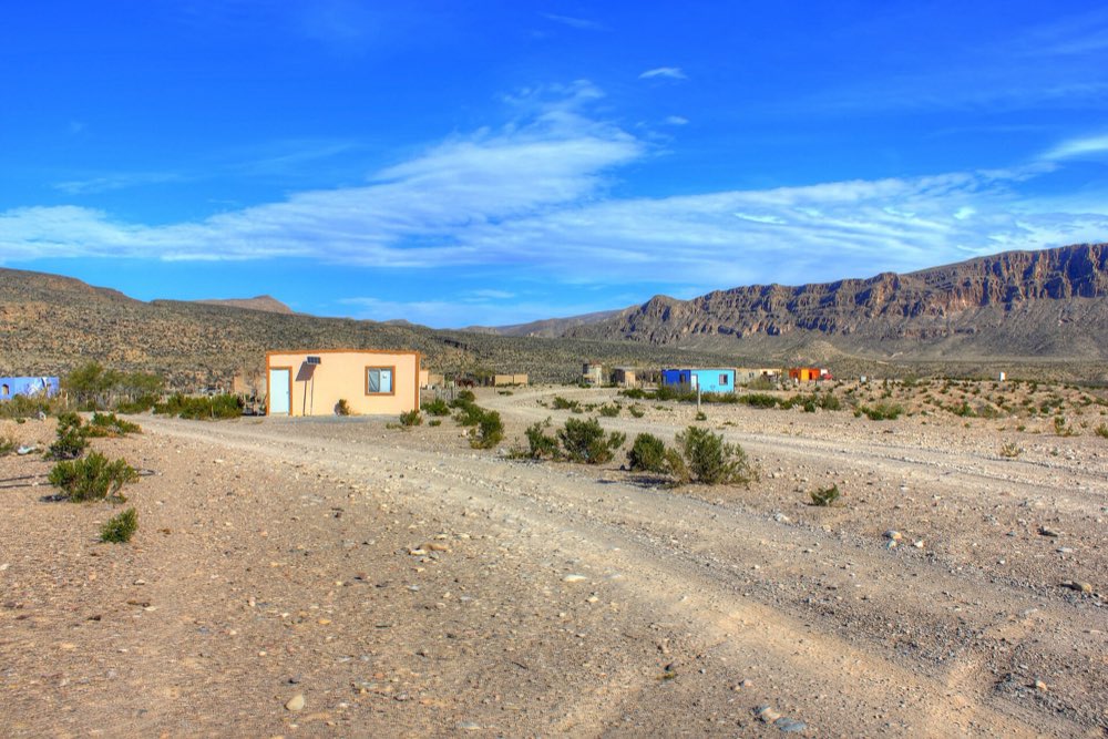 A dirt road leading past a few small houses in Boquillas del Carmen, with the long cliffs of the Sierra del Carmen under a deep blue sky