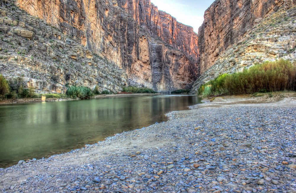 The Rio Grande at the mouth of Santa Elena Canyon between sheer limestone walls lit orange, with a pebble bar and green cane along the bank