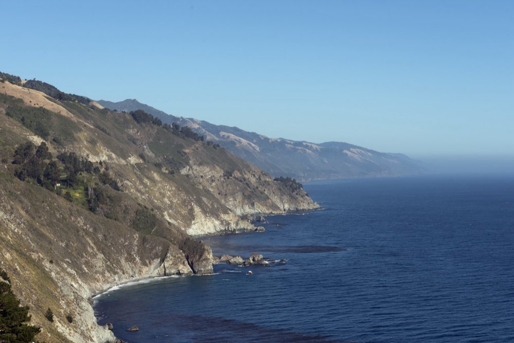 Steep golden and green Big Sur hillsides dropping straight into a deep blue Pacific, with headland after headland fading south into haze