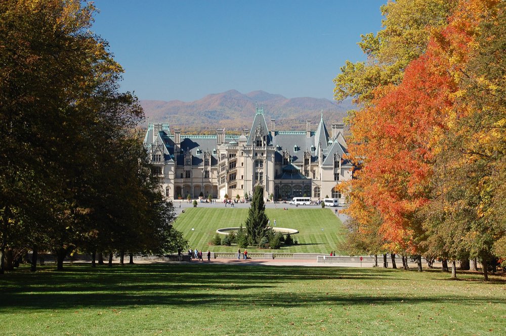Biltmore House seen down a long lawn between autumn trees in orange and gold, with blue mountains behind