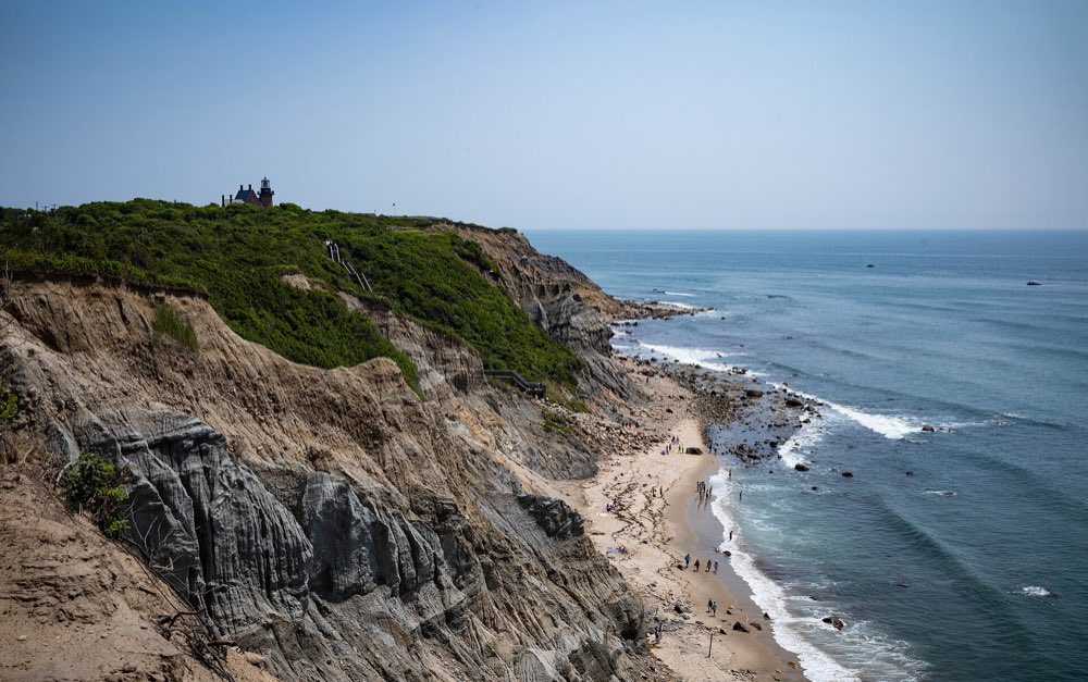 Tall eroded clay cliffs covered in green scrub drop to a narrow sandy beach and blue sea, with a brick lighthouse small on the clifftop
