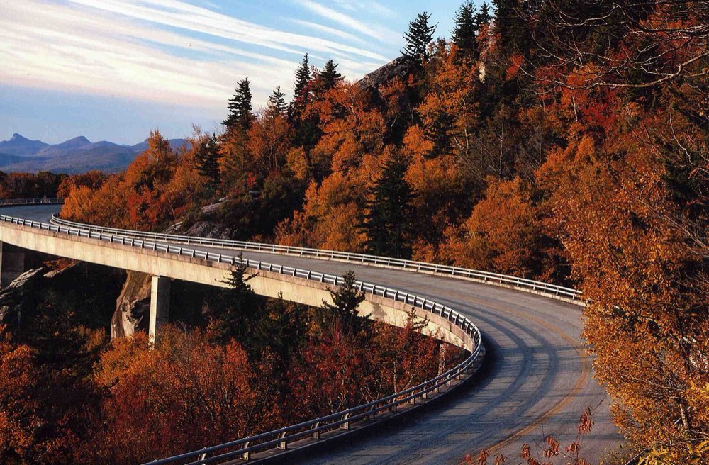 The curving concrete deck of the Linn Cove Viaduct hugging the side of Grandfather Mountain, surrounded by orange and red autumn trees, with blue mountains on the horizon