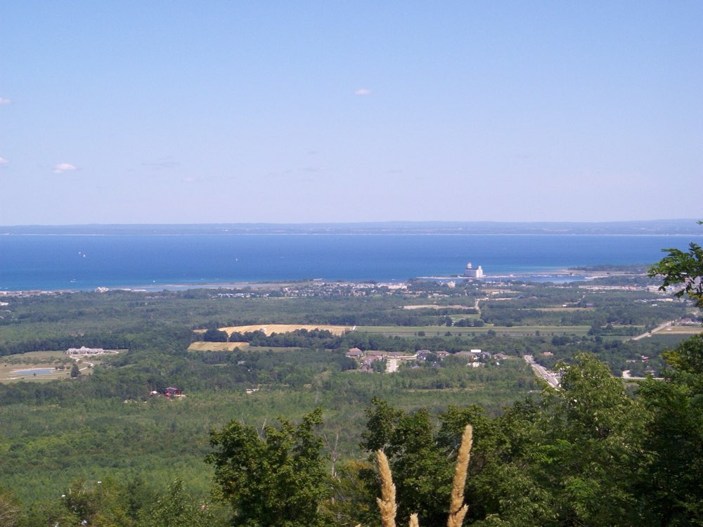 View from the top of Blue Mountain over forest and farmland to the blue water of Georgian Bay, with Collingwood's grain elevator on the shore