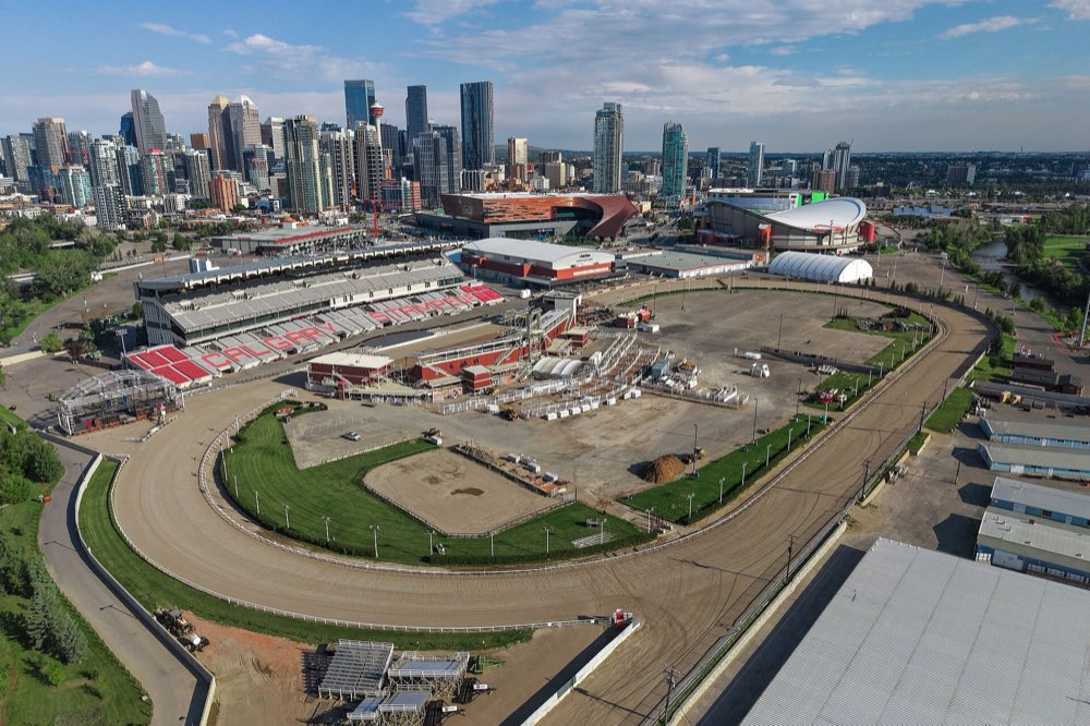 Aerial view of Stampede Park on a summer evening: the oval racetrack and grandstand of GMC Stadium in front, the curved roof of the Saddledome behind, and downtown Calgary's towers on the skyline