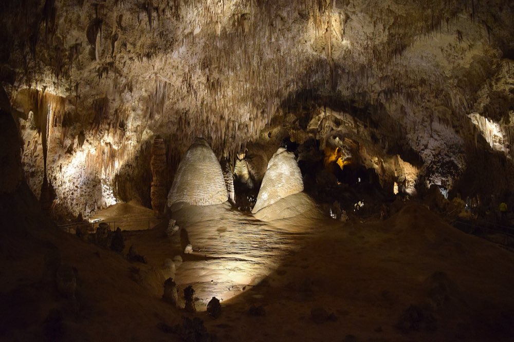 Two pale, rounded stalagmites lit in a vast dark cave chamber, with formations hanging from the ceiling and a lit trail winding between them
