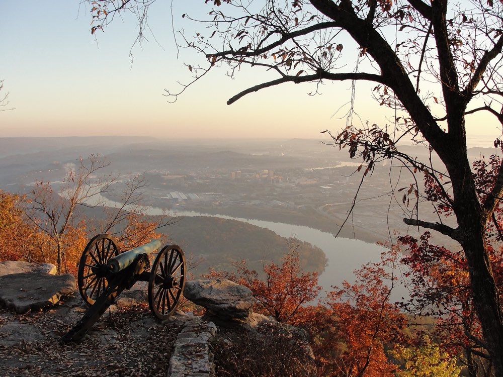 A Civil War cannon on a rocky ledge at Point Park in fall, with orange leaves and bare branches framing the Tennessee River curving around Moccasin Bend and the hazy city of Chattanooga below