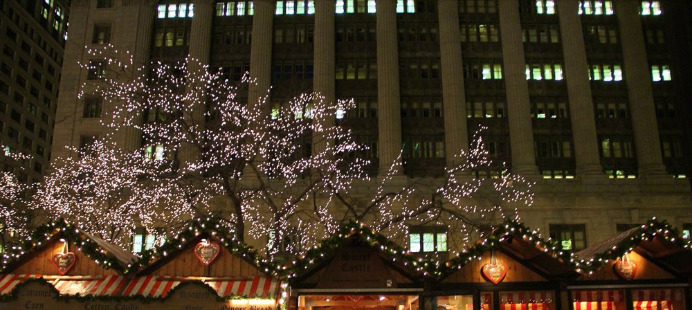 Wooden market stalls trimmed with garland and gingerbread hearts at night, under bare trees wrapped in white lights in front of a columned office tower