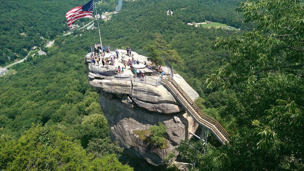 The flag-topped granite pillar of Chimney Rock with its stairway, seen from above, rising out of a forested valley