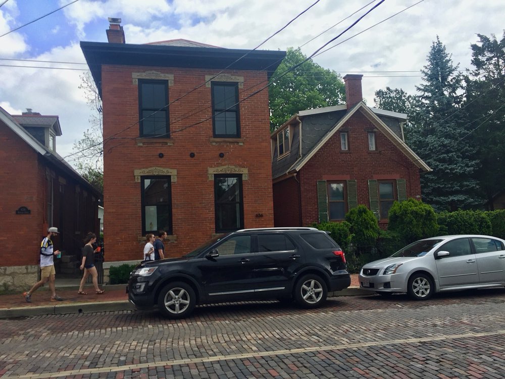 A red brick Italianate house with tall windows and stone lintels on a brick-paved street in German Village, with a cottage and green trees beside it