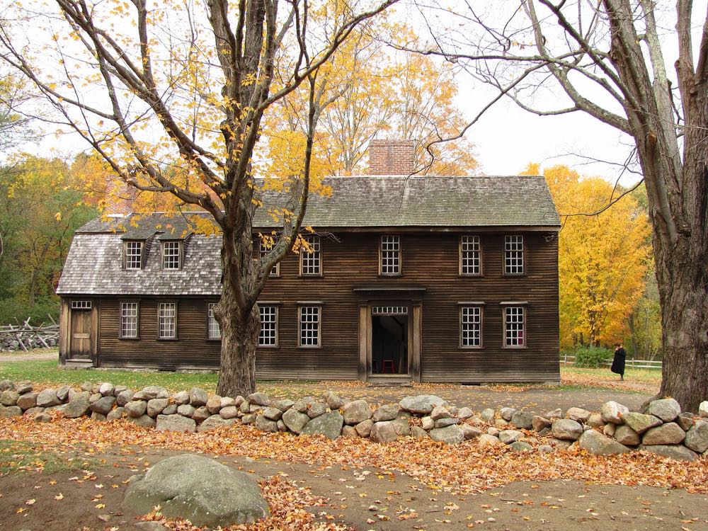 A dark-brown colonial tavern with a gray shingle roof behind a low fieldstone wall, framed by bare and golden autumn trees and fallen leaves