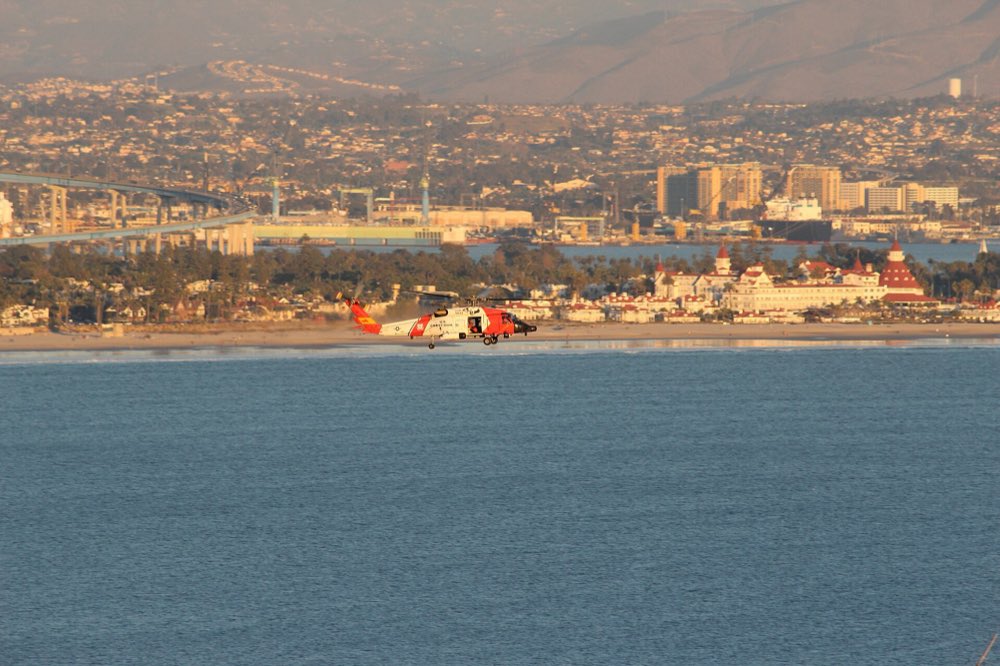 Looking across the blue mouth of San Diego Bay from Point Loma to Coronado, with the red-roofed Hotel del Coronado on the shore, a Coast Guard helicopter low over the water and the city and hills behind in warm evening light