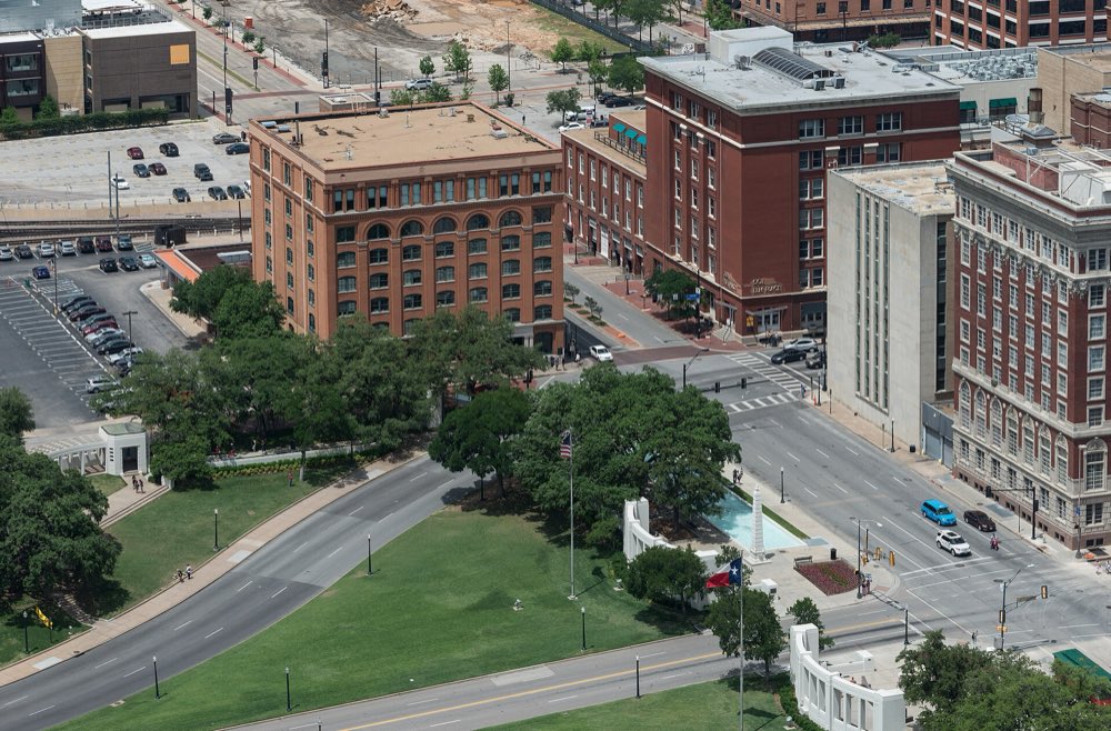 Elevated view of Dealey Plaza: the red-brick former Texas School Book Depository above the trees, Elm Street curving past green lawns and the white memorial structures