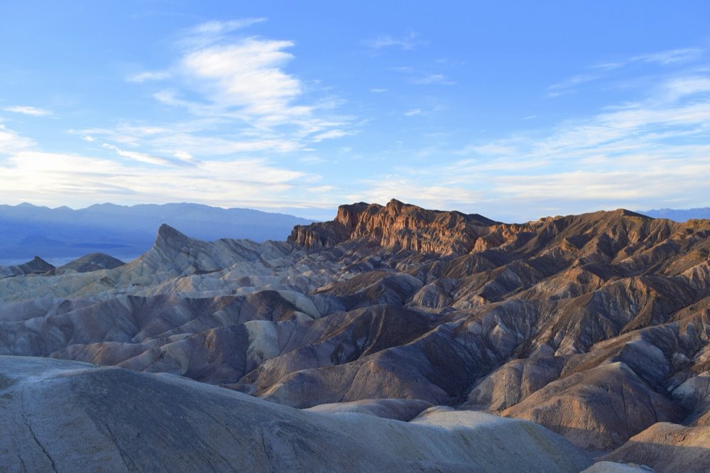 Rippled gray and tan badlands below the sunlit golden fin of Manly Beacon at Zabriskie Point, with blue mountains and wispy clouds beyond