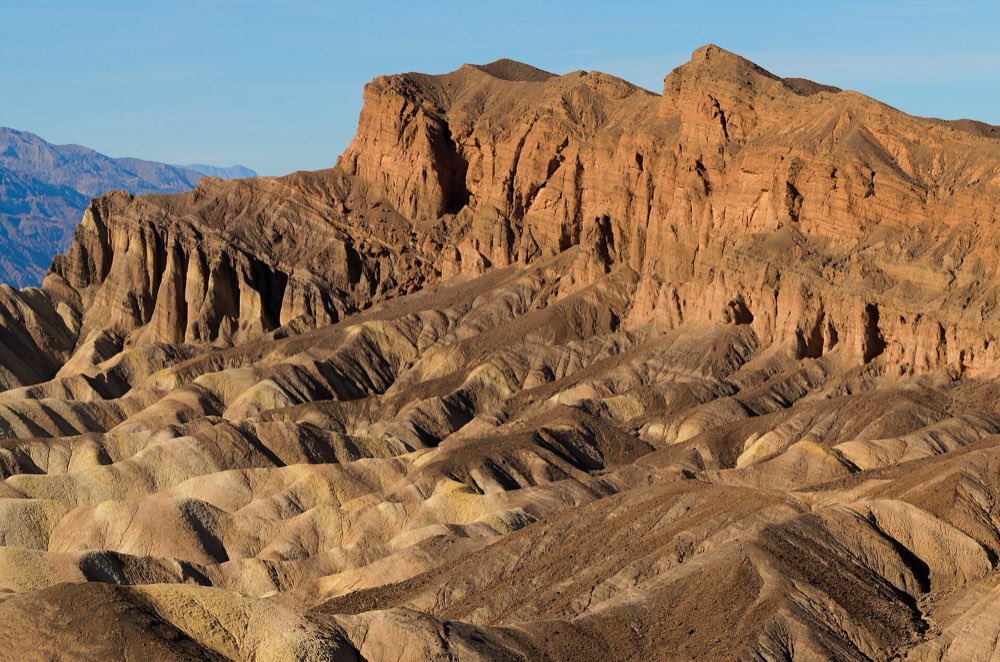 Golden eroded badlands in the foreground below a tall, sunlit ochre rock face at Zabriskie Point, with blue-gray mountains in the distance under a clear sky