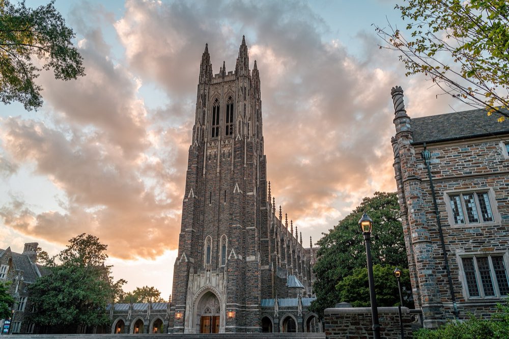 The tall stone Gothic tower of Duke Chapel against a sunset sky of pink and gold clouds, framed by trees and campus buildings