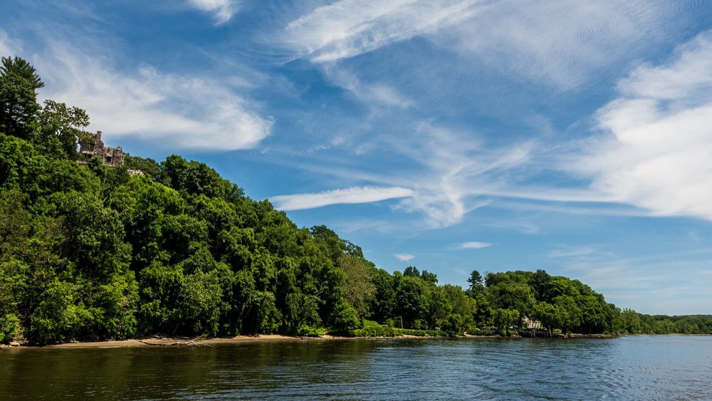 Wooded bluffs above the calm Connecticut River under a blue sky with high cloud, with Gillette Castle's stone towers just visible among the trees at the top left