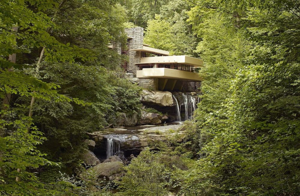 Fallingwater's cream-colored concrete terraces and stone chimney rising above a waterfall on Bear Run, framed by dense green summer forest