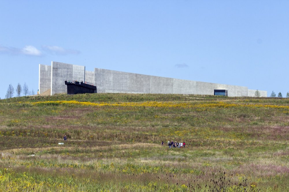 The long, pale concrete wall of the Flight 93 National Memorial visitor center along a hilltop, above a broad meadow of grasses and yellow wildflowers under a clear sky