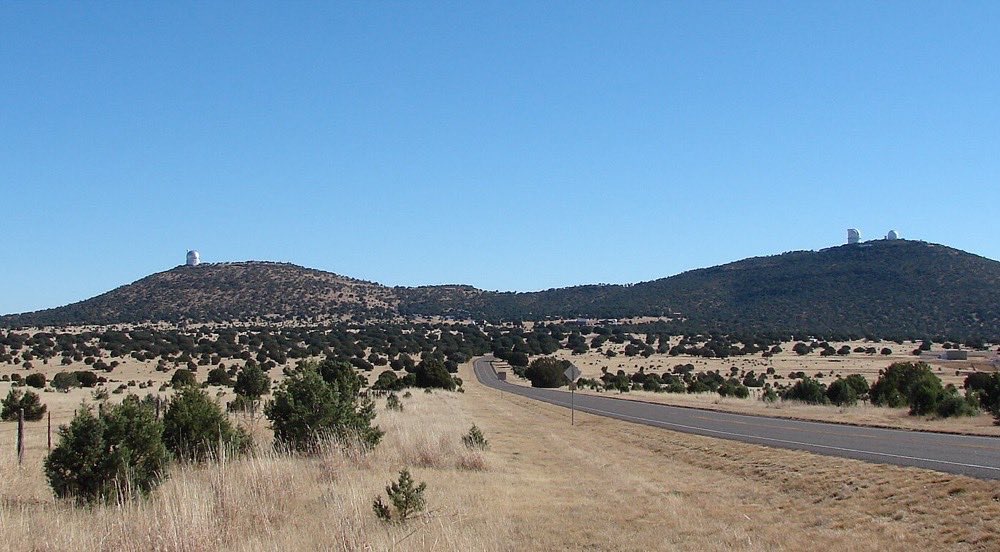 Two dry, rounded mountains with white observatory domes on their summits, seen across grassland and juniper from a two-lane highway under a clear blue sky