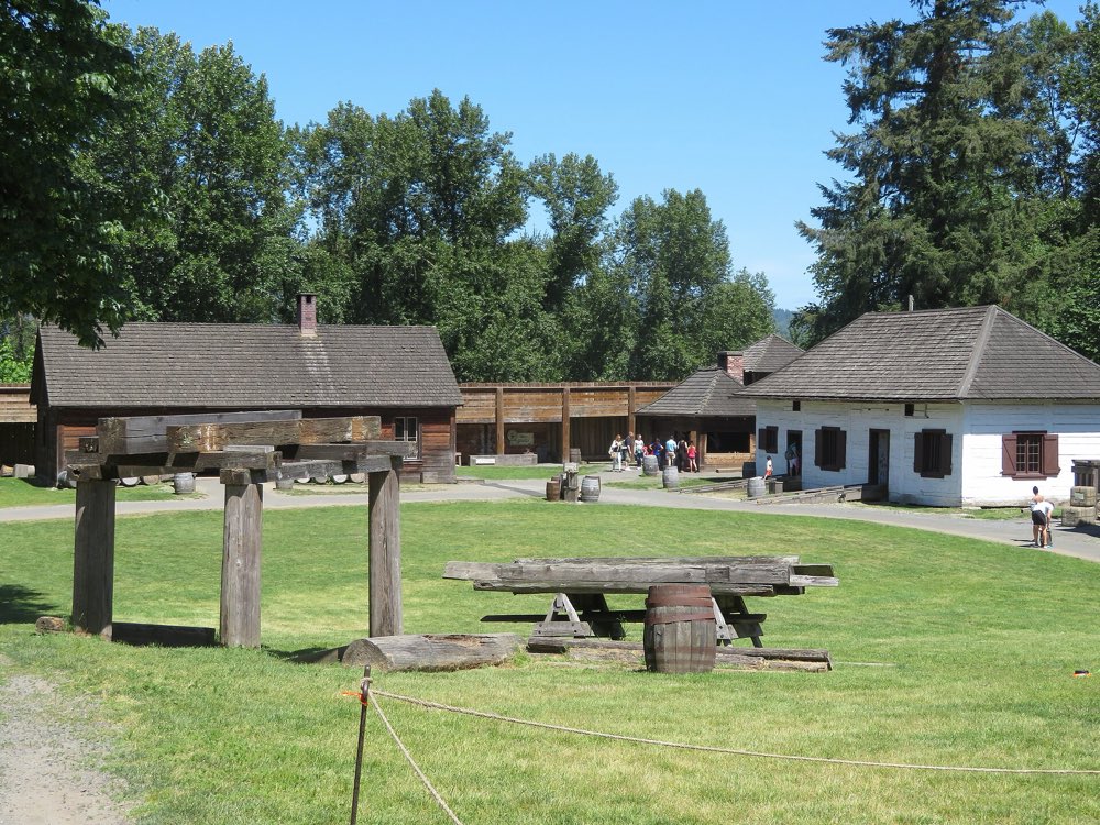 Inside the timber palisade at Fort Langley: a wooden saw pit and log benches on a sunny lawn, with a squared-log storehouse and a white clapboard building behind under tall trees