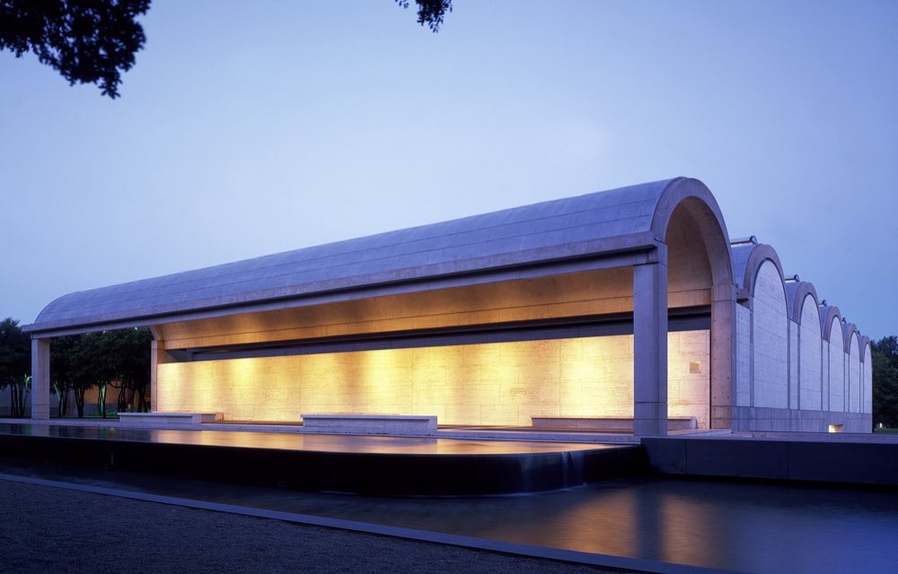 The Kimbell Art Museum's open, barrel-vaulted portico glowing warm at dusk above a long reflecting pool, with tree branches framing the sky