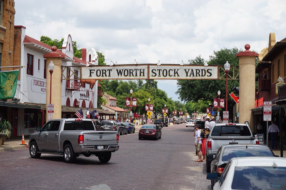 The Fort Worth Stock Yards sign arching over brick-paved Exchange Avenue, with western storefronts and parked pickups on both sides