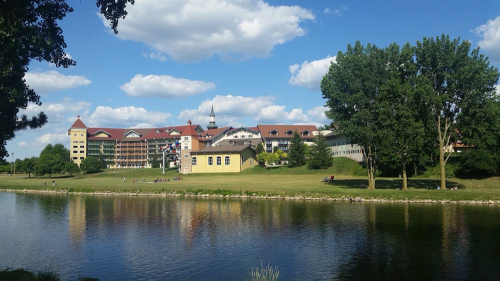 The long Bavarian-style Bavarian Inn Lodge with red roofs and a white clock tower, seen across the calm Cass River and a broad green lawn under a summer sky with white clouds