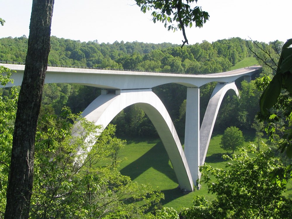 The white concrete Double Arch Bridge carrying the Natchez Trace Parkway high over a green, wooded hollow, framed by leafy branches
