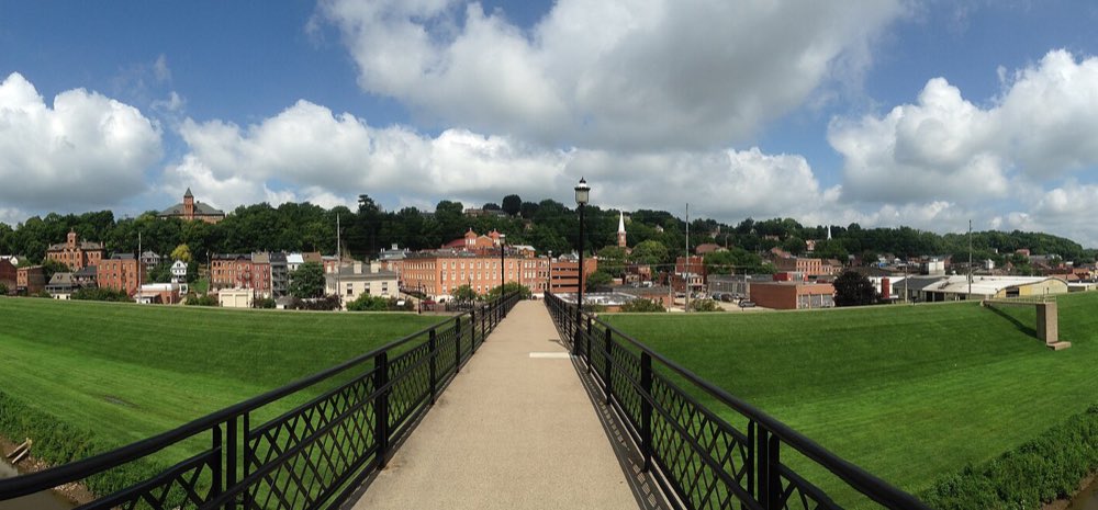 A footbridge with black iron railings crossing the grassy levee toward downtown Galena, where brick buildings and church steeples climb a wooded hillside under puffy white clouds