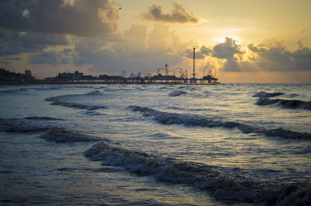 Low waves rolling onto a Galveston beach at sunrise, with the Pleasure Pier's roller coaster, tower and Ferris wheel silhouetted on the horizon under a golden, cloudy sky