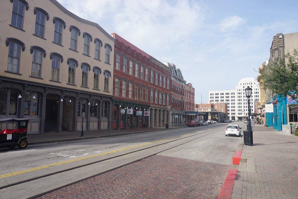 A brick-paved stretch of The Strand with rail trolley tracks, three- and four-story Victorian commercial buildings with arched windows, street lamps and a parked open trolley car