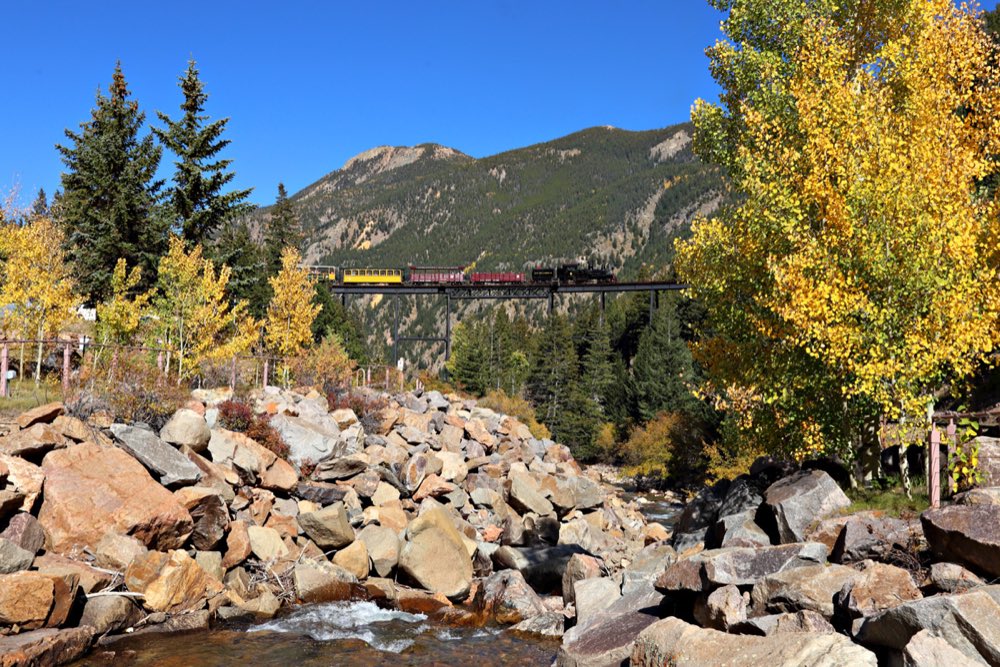 A steam train with yellow and red cars crossing a high steel trestle above a rocky creek, framed by golden aspens and a mountain under a blue sky