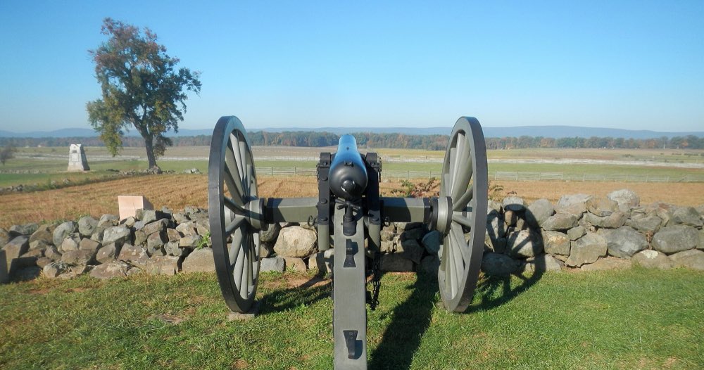 A Civil War-era cannon behind a low stone wall on Cemetery Ridge, aimed across a mile of open fields and fences toward a distant tree line and blue ridges under a clear sky