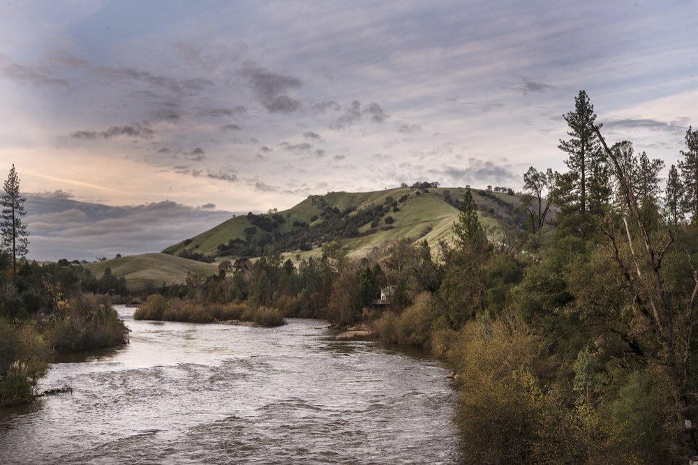 The South Fork of the American River curving past oak and pine woods below green hills at Coloma, under a cloudy evening sky