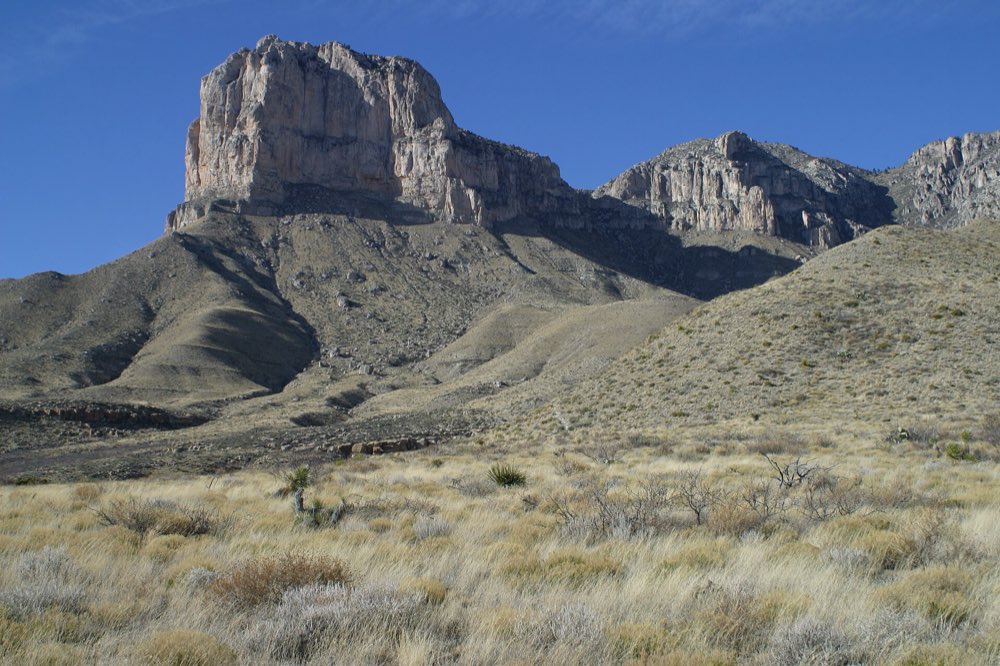 The pale limestone cliff of El Capitan rising above sloping desert hills, with dry golden grass in the foreground under a deep blue sky