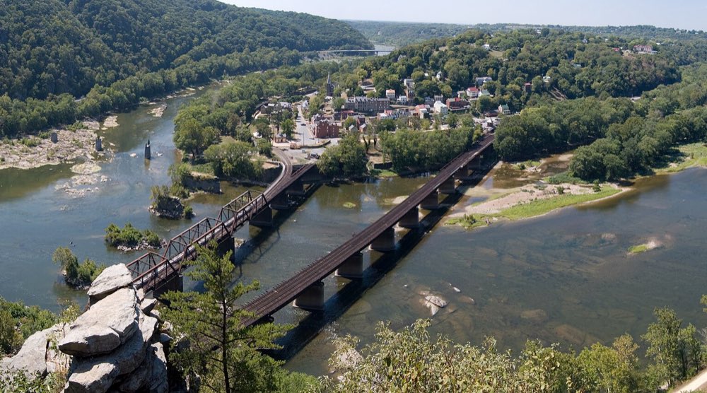 Harpers Ferry seen from a rocky cliff high above the Potomac: two railroad bridges cross the wide river toward the old brick town on its wooded point, with the Shenandoah curving in from the left