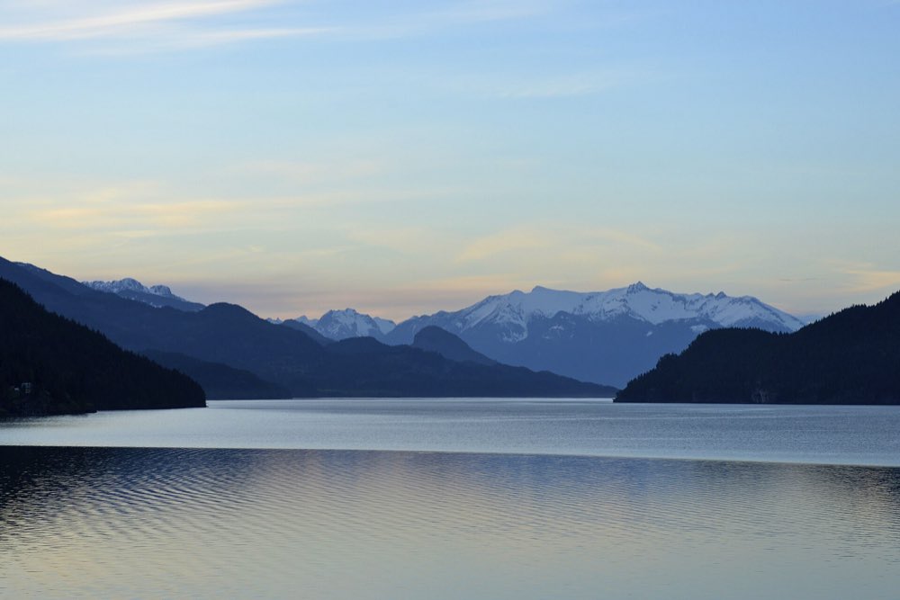 Harrison Lake at dusk, its still water reflecting a pale sky, with dark forested ridges on both sides and snow-capped peaks at the far end