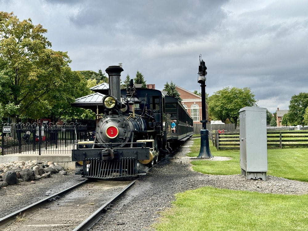 A small black steam locomotive with a red number 1 on its smokebox waiting on the track at a Greenfield Village station, with green lawns and brick buildings behind