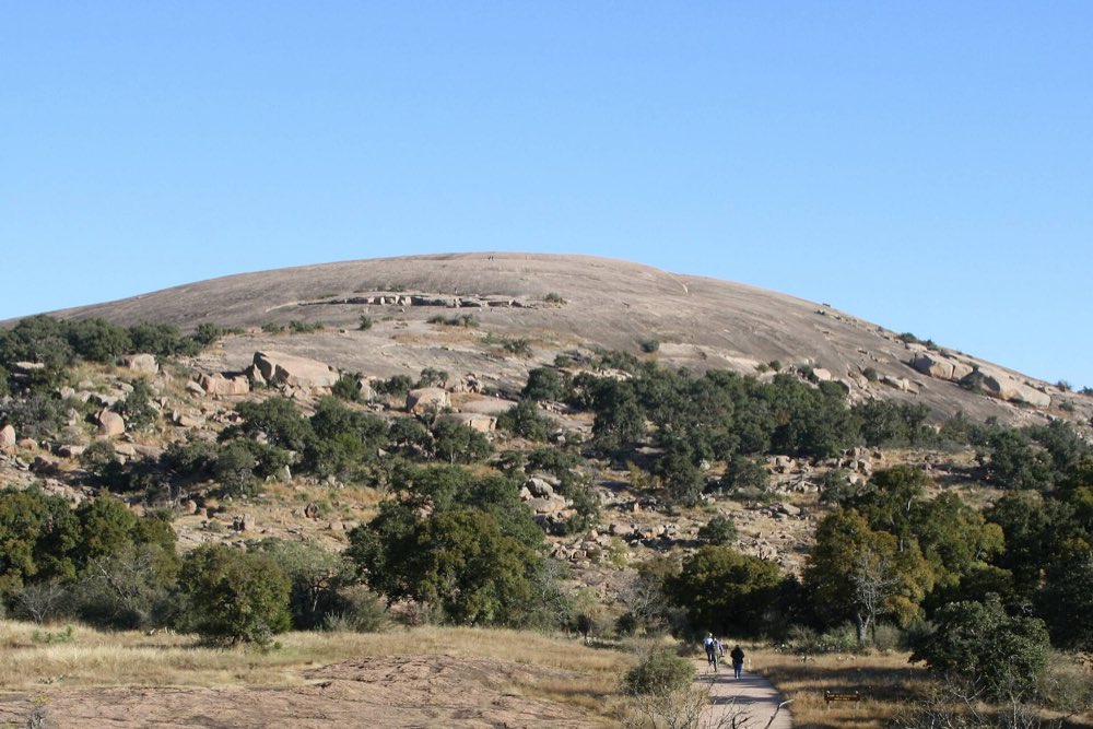 The bare pink granite dome of Enchanted Rock rising above oak scrub and boulders, with two hikers small on a path in the foreground under a clear blue sky