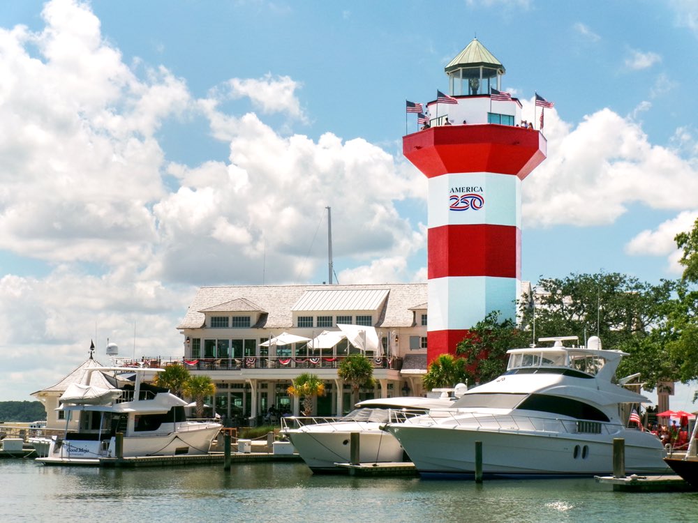 The red-and-white striped Harbour Town Lighthouse rising above yachts moored in the marina under a bright, cloudy summer sky