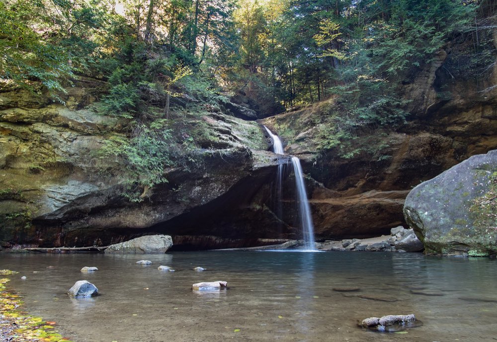 A thin waterfall drops over a dark sandstone ledge into a wide, still pool in the Old Man's Cave gorge, with hemlocks and a large boulder on the bank